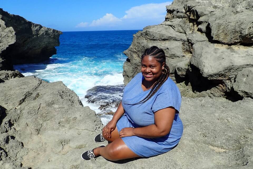 Diedre McLeod from travel blog Diedre in Wanderland. A curvy black woman in big cornrow braids sits at the edge of a beachy cliff, smiling and looking directly into camera. The waves are crashing into the cliffs in the background.