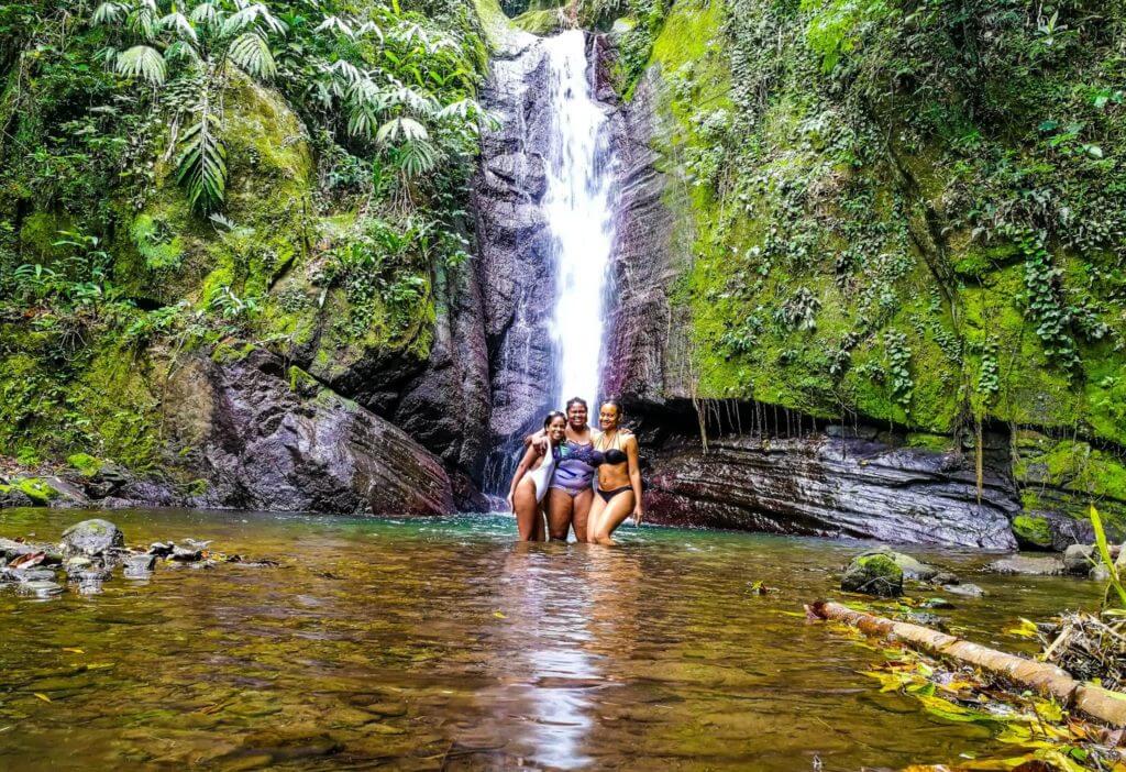 three black women friends pose standing in front of Falling Edge waterfall with clear turquoise water in Jamaica