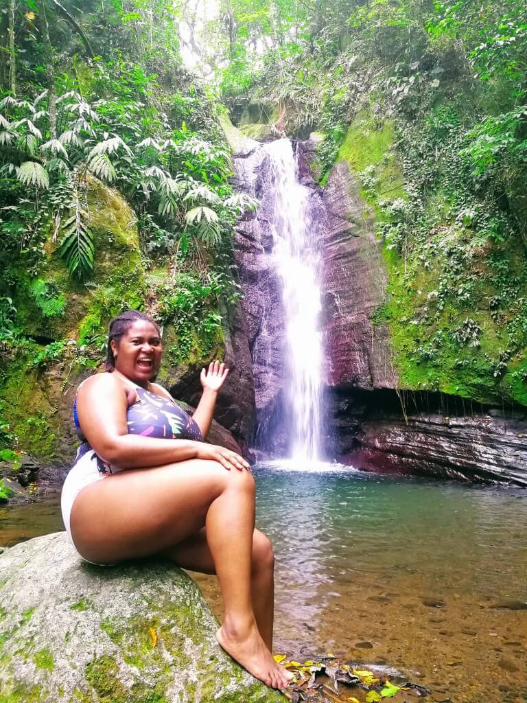 Happy Black woman in swimsuit sitting on a rock gesturing to falling edge waterfall and hidden gem in Kingston, Jamaica.