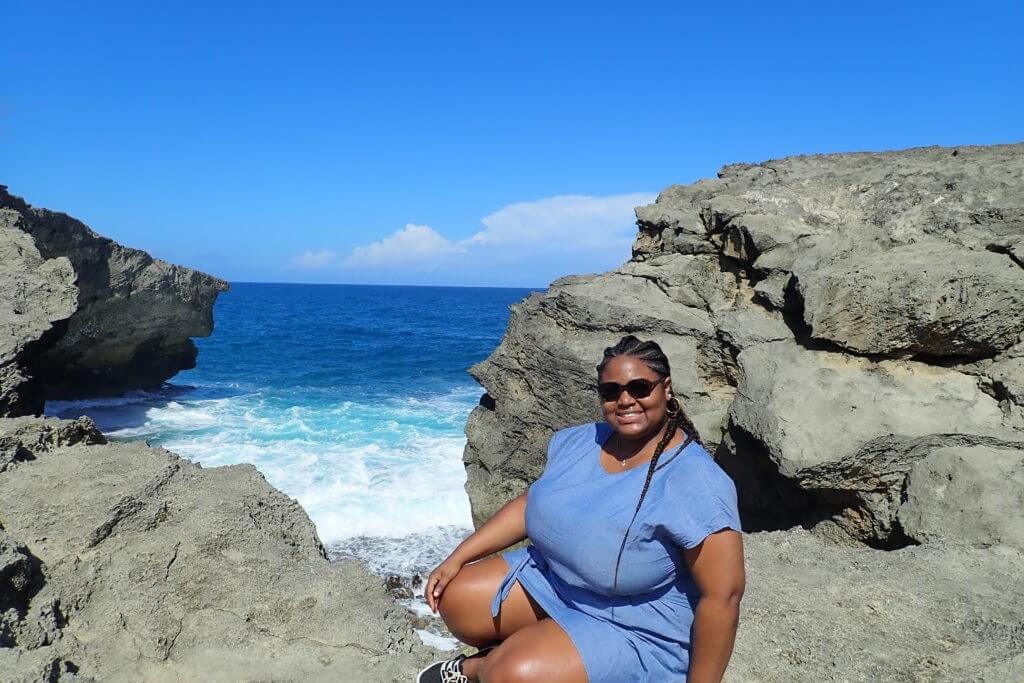 A smiling black curvy female traveler sits at the edge of a cliff in puerto rico, with deep blue beach waves in the background