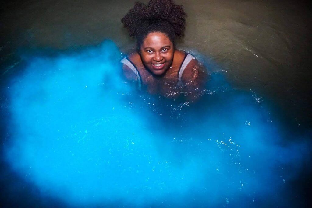 Black woman swims in bioluminescent lagoon in jamaica. She is smiling and looking up into the camera. the water is glowing a bright neon blue all around her.