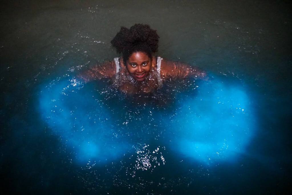 Curvy black woman swimming in a luminous lagoon with waters that glow bright blue when disturbed.