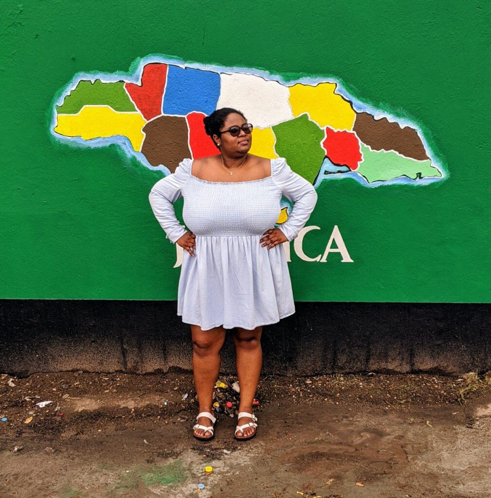 Black curvy woman poses confidently in front of a Jamaica Map mural downtown jamaica
