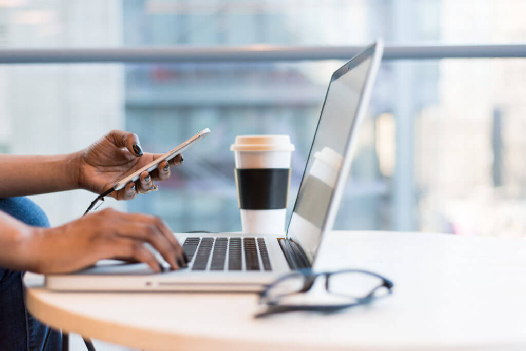 hands of a black woman typing on a laptop while holding a cell phone in the left hand. Photo is of an office or work from home space