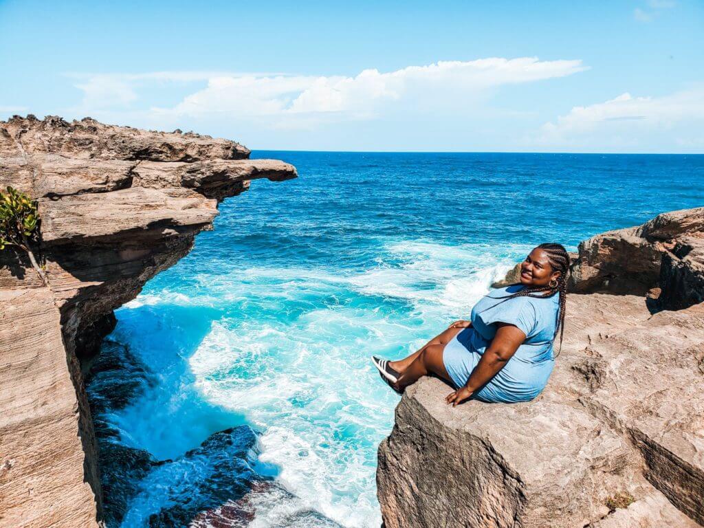 Curvy black woman sitting at the edge of a sea cliff, smiling and lookin from behind her shoulders