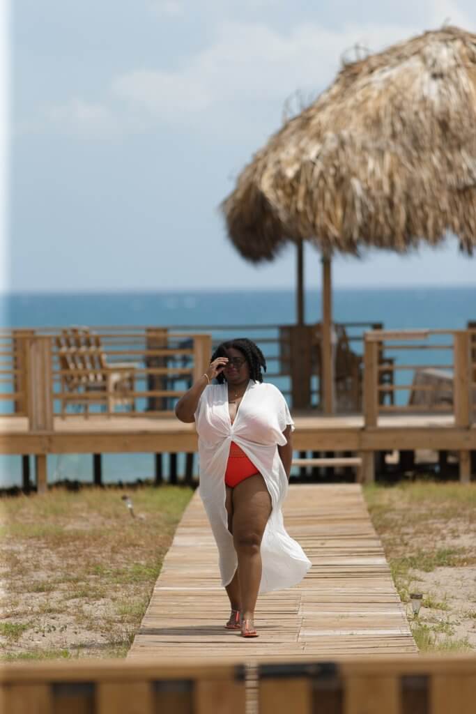 Plus size Black woman in orange swimsuit and white coverup walks along the boardwalk at Turtles' Nest Villa.