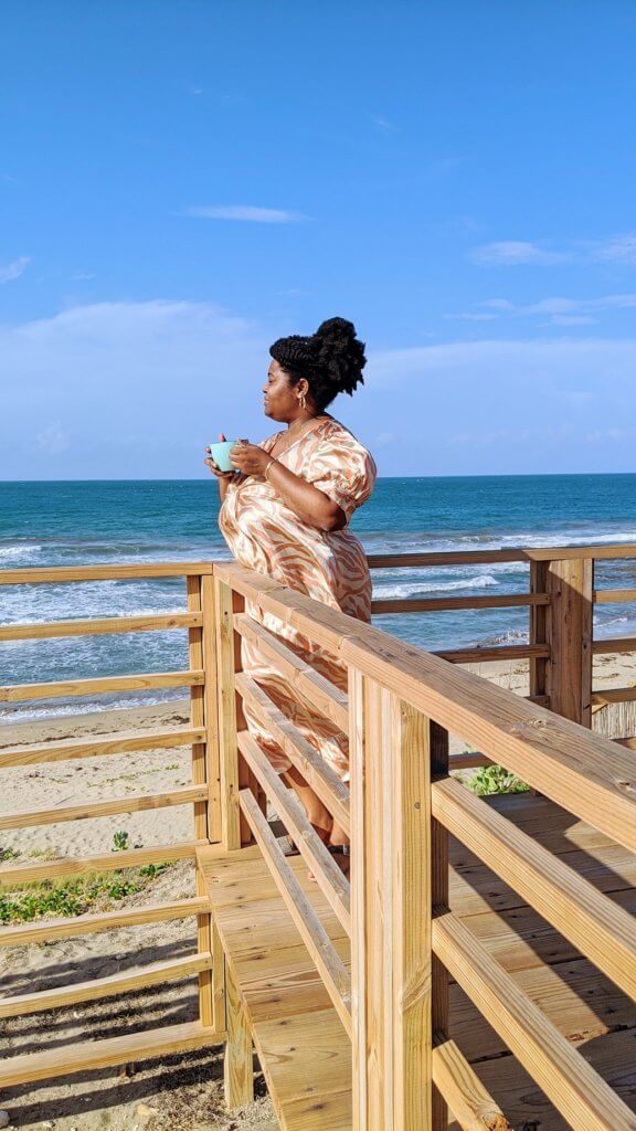 Black curvy women in long flowing dress have a cup of tea on the wooden deck at Turtles' Nest Villa