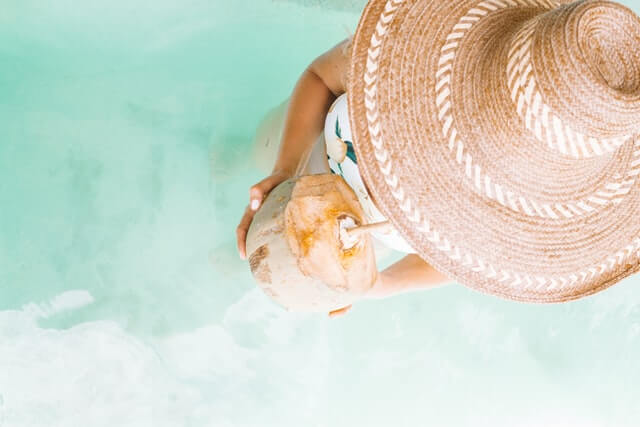 overhead shot of a woman standing in powdery blue sea water, wearing a broad hat and sipping a coconut