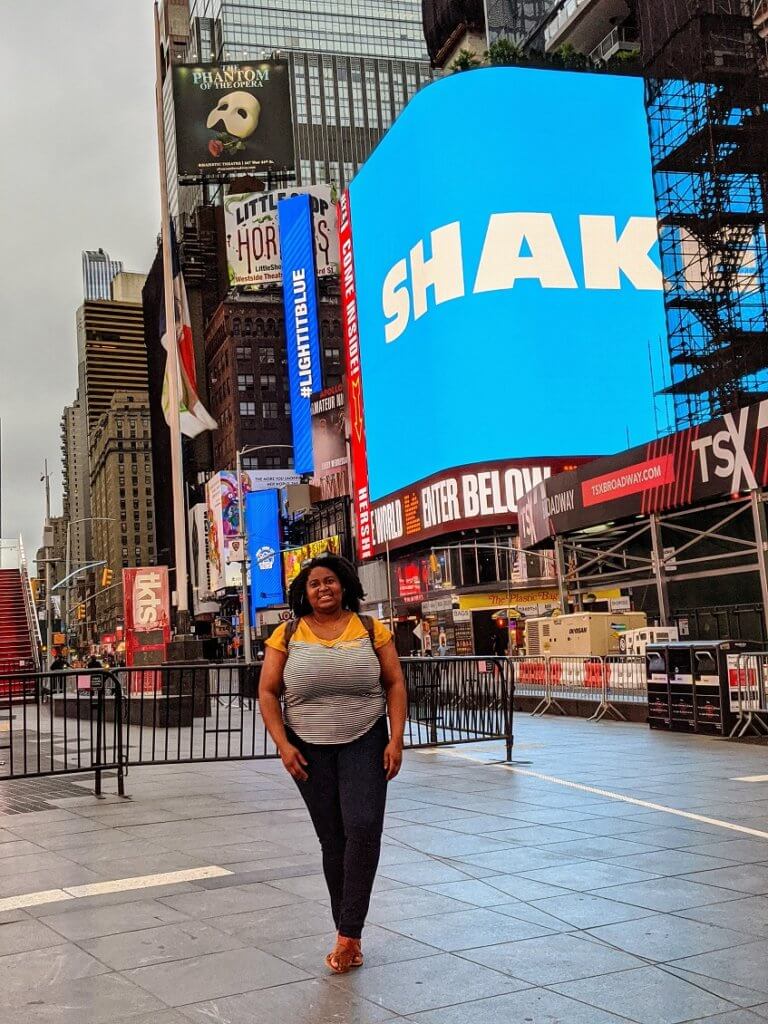 Plus size Black woman in a baby-tee with gold shoulders and black and white stripes. The Black woman has natural hair worn in a curly twist out. She stands happil in the middle of Times Square which is empty because of covid-19 shutdown