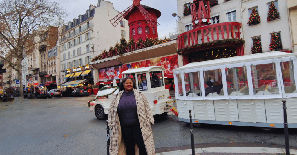 smiling Black woman at the Moulin Rouge in Paris, France during Fall/Autumn season. The woman is wearing a long beige and black checkered coat with a dark grey sweater, black leggings, and black calf boots.